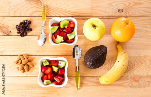 Two bowls ready for a healthy break with fresh fruit cutted for a salad. Strawberries and kiwi. Orange and banana. Wood background with dried fruit and avocado. No people