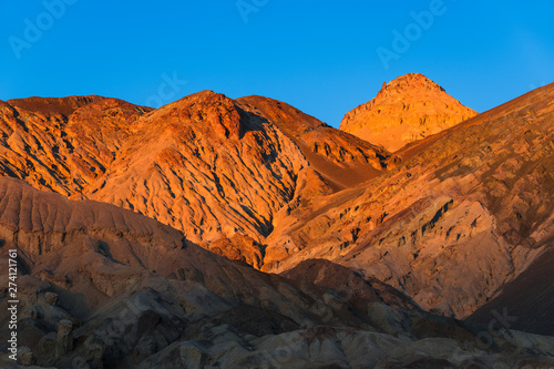 Barren desert mountains and a high peak glow in the golden hour of sunset under a perfect blue sky - Death Valley National Park, California