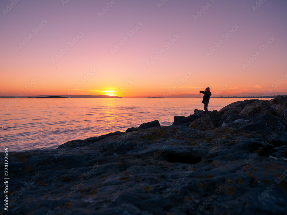 Silhouette of teenager girl pointing at sun set over ocean, Galway bay, Ireland. Concept, childhood, adventure, excitement. Silhouette of teenager girl pointing at sun set over ocean, Galway bay, Ireland. Concept, childhood, adventure, excitement.