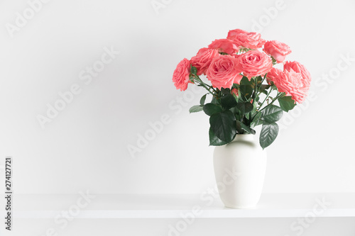 Roses flowers of coral color in vase on shelf against white wall. Space for text © prime1001