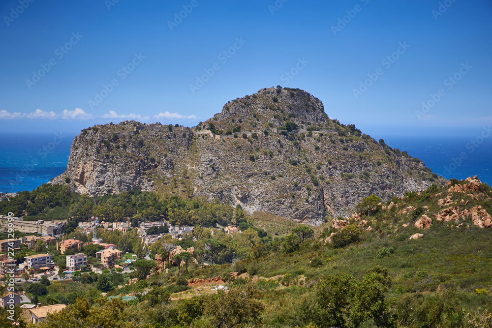 Landscape of historic italian city Cefalu in the island Sicily with ruined mediaeval fortress or castle on the top of the mountain which protected city agains pirate raids and blue sea in background.