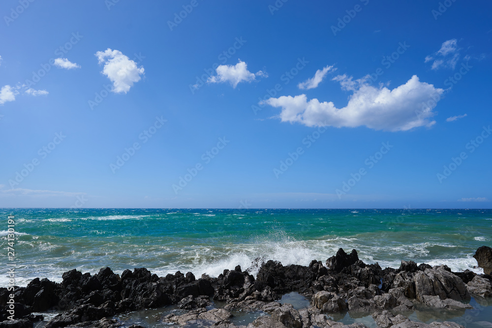 Landscape Picture of coastline in windy sunny day with black lava rocks and blue rough sea, wawes and sky with white clouds. Taken in italian island sicily in volcanic area of mediterranean sea.
