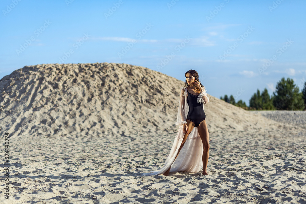Attractive brunette woman in black body and translucent beach cover up posing on sandy beach at sunset and looking away. Fashion outdoor shot in the summertime. Attractive brunette woman in black body and translucent beach cover up posing on sandy beach at sunset and looking away. Fashion outdoor shot in the summertime.