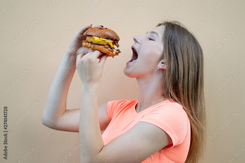 attractive teen girl stands against the orange wall in an orange T-shirt and holds a big burger in her hand, she opens her mouth wide and eats it attractive teen girl stands against the orange wall in an orange T-shirt and holds a big burger in her hand, she opens her mouth wide and eats it