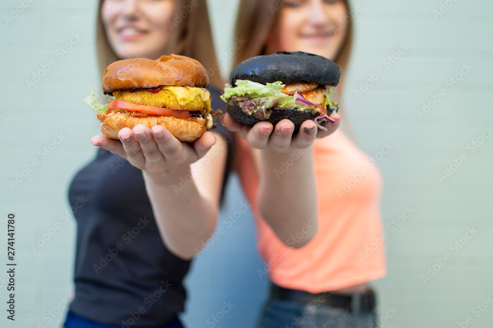 young beautiful girls-students stand near the blue wall, hold burgers and smile, they are happy and show cheeseburgers on camera young beautiful girls-students stand near the blue wall, hold burgers and smile, they are happy and show cheeseburgers on camera