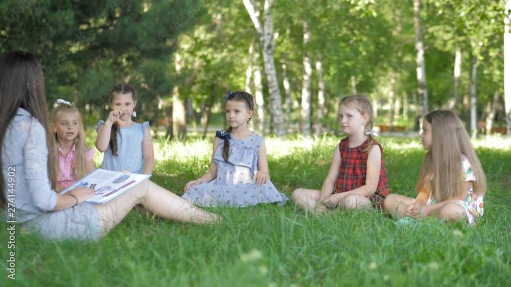 Children and education, young woman at work as educator reading book to boys and girls in park Children and education, young woman at work as educator reading book to boys and girls in park