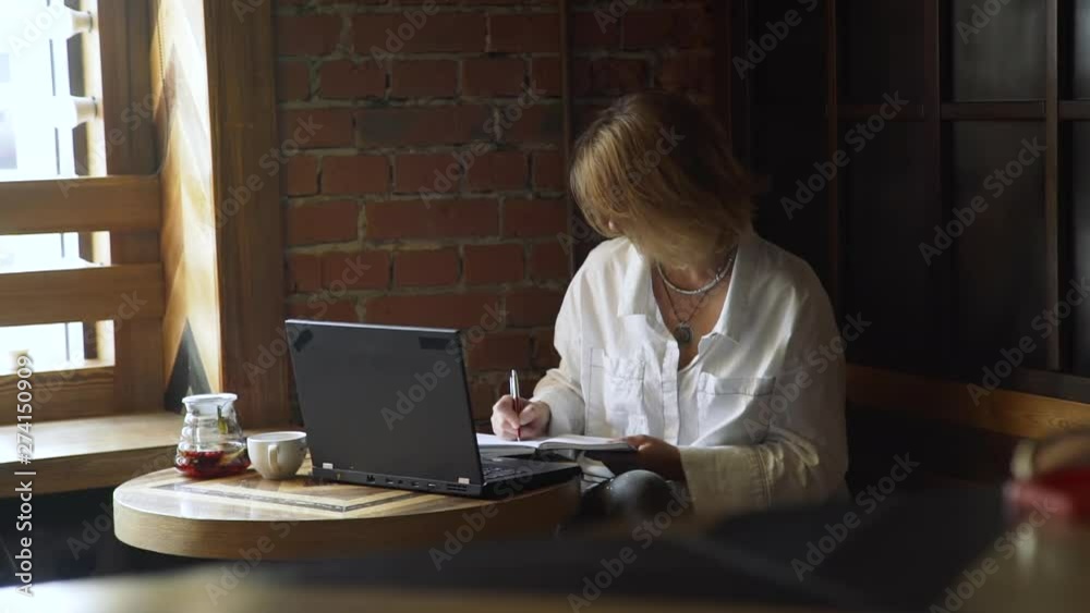 Middle Age Business Woman Working Remotely in a Coffee Shop in Slow Motion. She is Making Notes of Ideas in her Notebook. Freelance Work, Business People Concept Middle Age Business Woman Working Remotely in a Coffee Shop in Slow Motion. She is Making Notes of Ideas in her Notebook. Freelance Work, Business People Concept