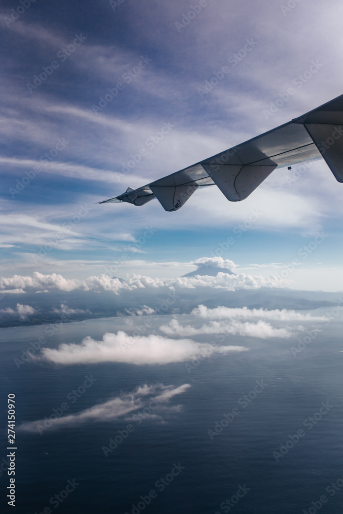 Fototapeta premium Plane window view with blue sky and beautiful clouds, sky. As seen through window of an aircraft. View of wing, ocean, volcano Agung & Nusa Penida. Airplane from island Bali to Labuan Bajo, Komodo.