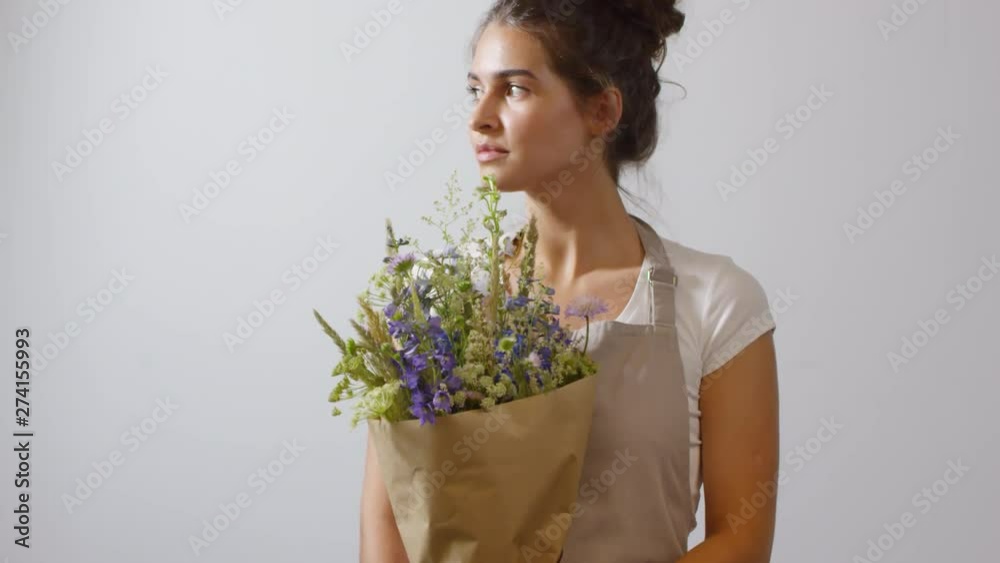 Tilting portrait shot of smiling beautiful dark-haired florist posing with her freshly arranged delicate wildflower bouquet wrapped in plain brown paper against beige-grey wall of her flower shop Tilting portrait shot of smiling beautiful dark-haired florist posing with her freshly arranged delicate wildflower bouquet wrapped in plain brown paper against beige-grey wall of her flower shop