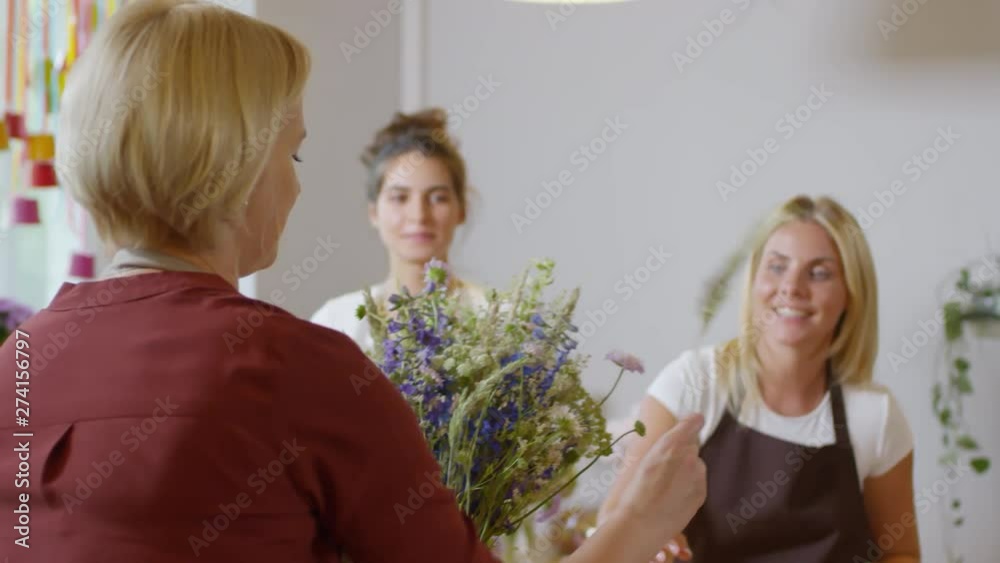 Medium shot of experienced florist, standing with her back to camera, giving demonstration for two young enthusiastic apprentices on how to arrange exquisite bouquet with wild flowers and dried grass