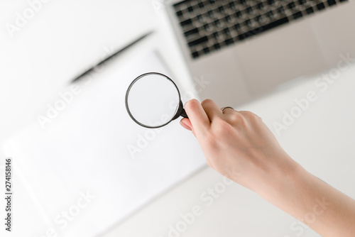 Hand with magnifying glass in focus, laptop and note book on background