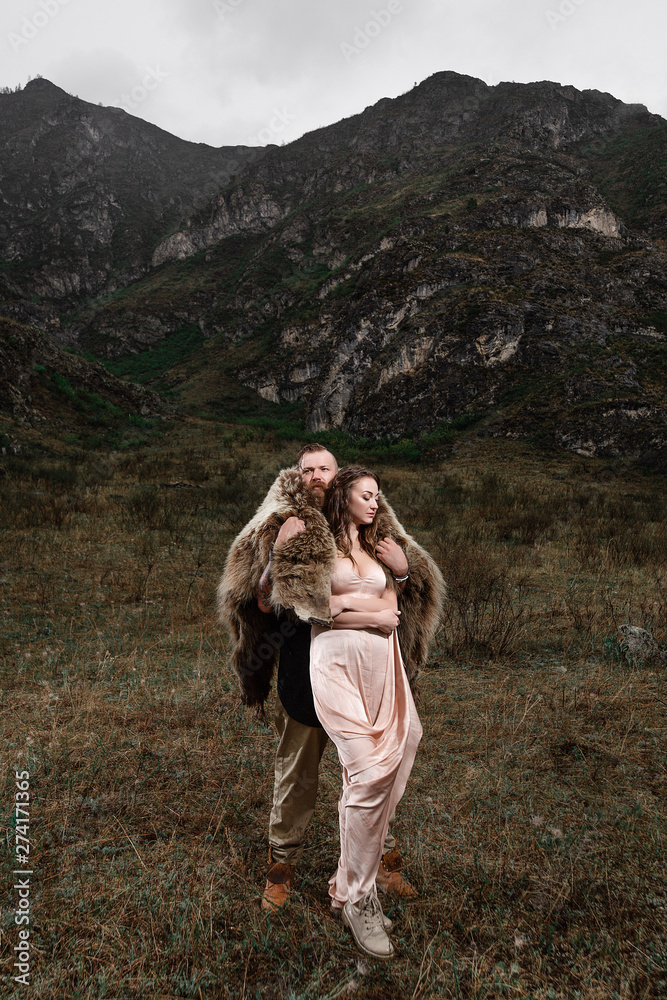 a serious and strict man with a beard and mustache, in a black shirt and a bear's skin, stands on a rock, a mountain. hugging a girl in a cream dress