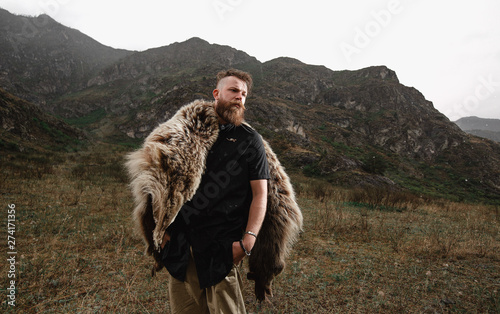 Bild auf Leinwand a serious and strict man with a beard and mustache in a black T-shirt and a bear skin stands at a cliff, a mountain