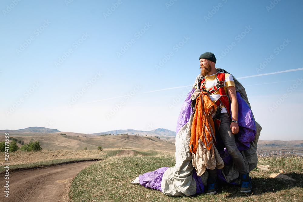 Fototapeta premium man with beard mustache striped white-yellow T-shirt and a green hat jeans with a red backpack. Carries a purple gray parachute on background of the mountains, a tattoo on his arm