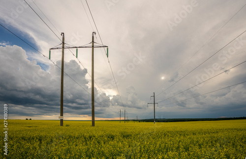 high-voltage power line in the field of blooming yellow rape flowers on the background of the sky with storm clouds