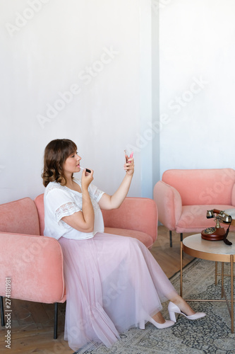 A woman sits on the sofa in the room and paints her lips looking at the reflection in the phone. Girl plus size in tutu skirt and white top. Self-care, beauty fashion concept.