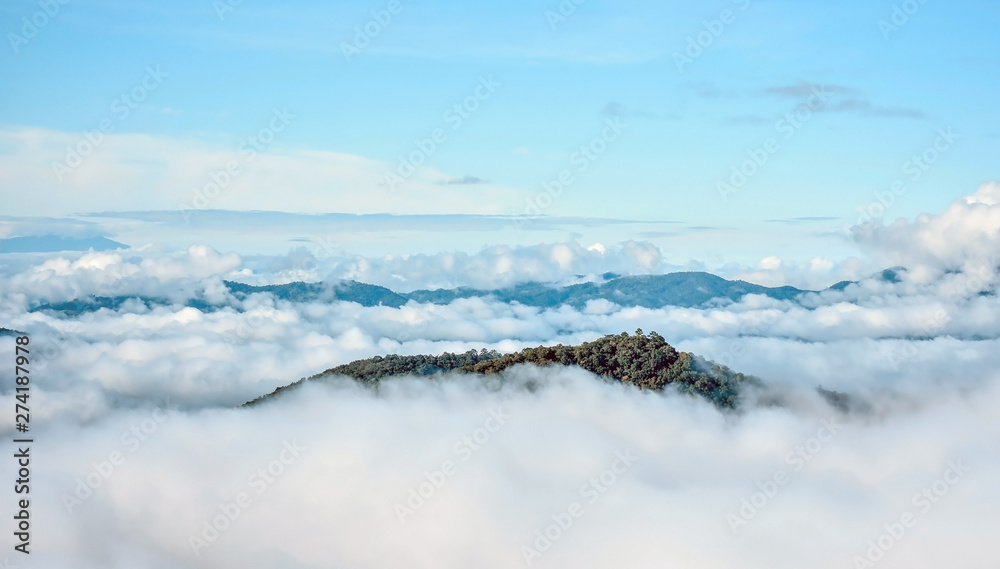 Blue mountain ridge emerges over vast sea of fog in the valley of northern Thailand. Thick green forest in foreground and morning clear blue sky in the background.