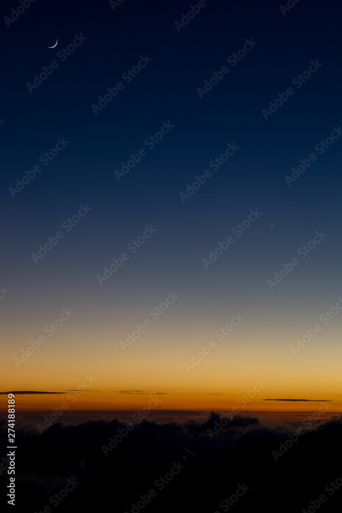 Crescent moon over the bright orange horizon lit by the last light of sunset at the Mauna Kea on Big Island, Hawaii, USA.