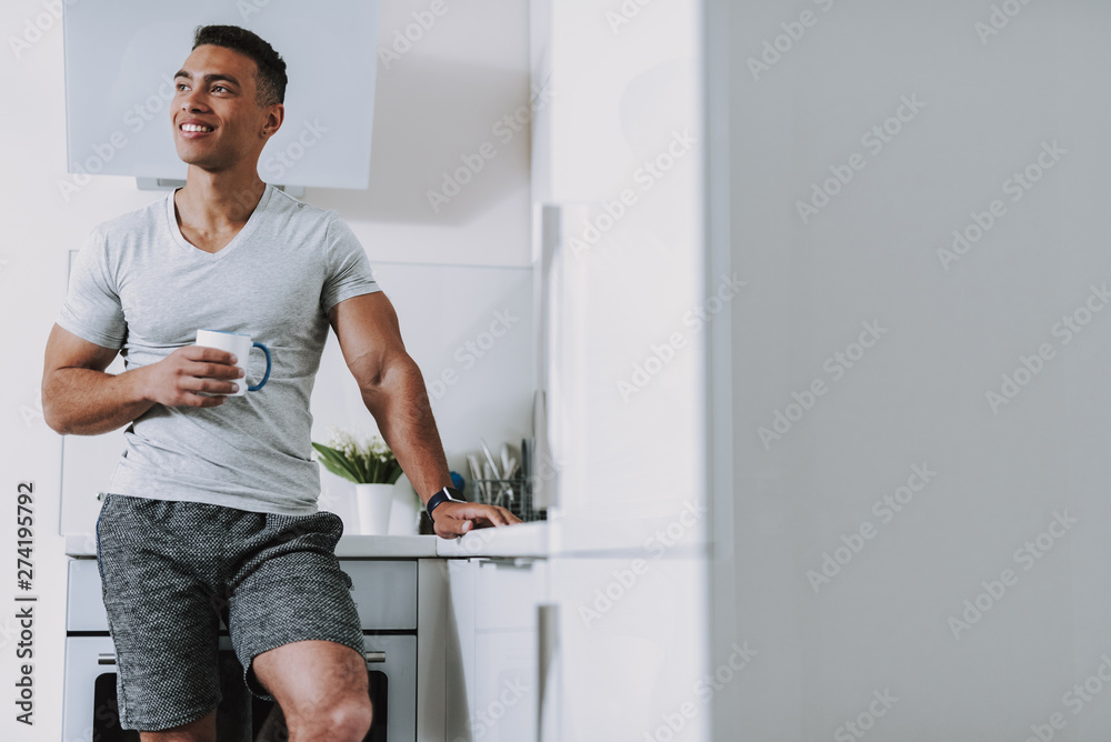 Cheerful young man is drinking hot beverage in kitchen