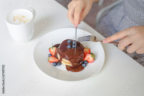 Close up of a women eats pancakes for breakfast at the kitchen table. Pancakes with chocolate and blueberry on the top and cup of coffe.
