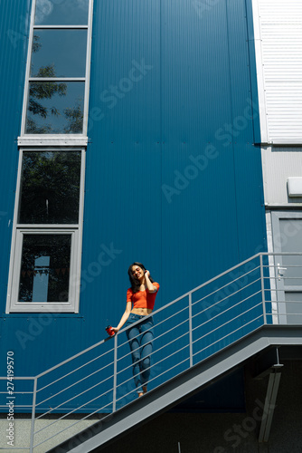 Charming young woman with cup of coffee standing on stairs against blue wall