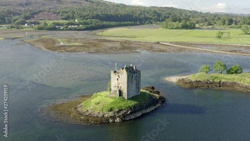 An aerial, close view of Castle Stalker on Loch Laich on a sunny morning. Flying left to right whilst ascending, with forests and fields behind the castle. photo