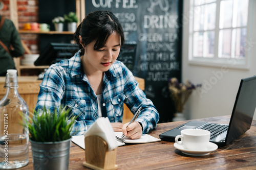 front view of young female freelancer in plaid shirt sitting at table in cafe bar working writing in notebook. laptop and cup of coffee. cute college girl student learning online notebook pc
