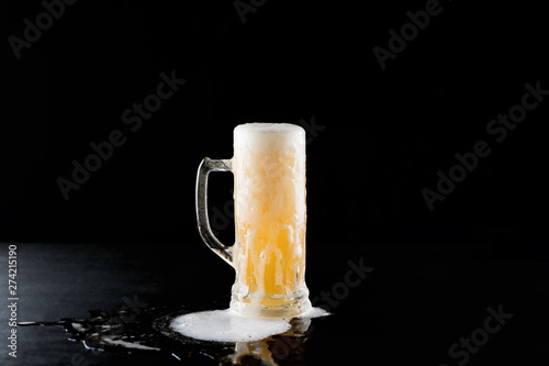 overflowing mug of fresh cold light beer with froth flowing on the table and beautiful spilled beer on a black background with reflection