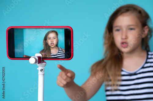Girl posing in front of phone camera for selfie. Focus o smartphone screen fixed on tripod. Recording homemade video. Happy smiling child. Cute kid pointing up. Simple wallpaper, background