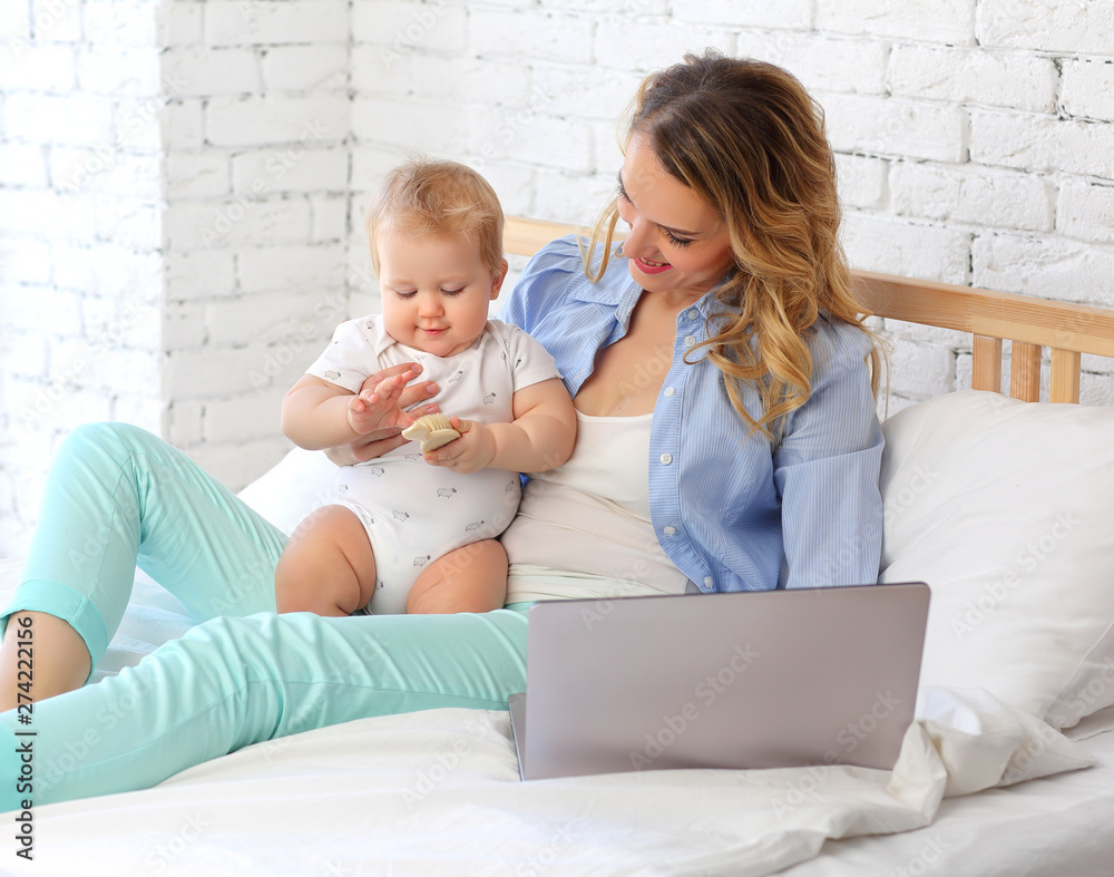 A young mother works at a laptop on the bed and hugs her 9-month-old baby A young mother works at a laptop on the bed and hugs her 9-month-old baby