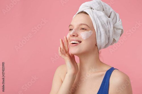 Photo of young nice joyful lady with natural beauty with a towel on her head after shower, stands over pink background and puts on face cream, looks away.