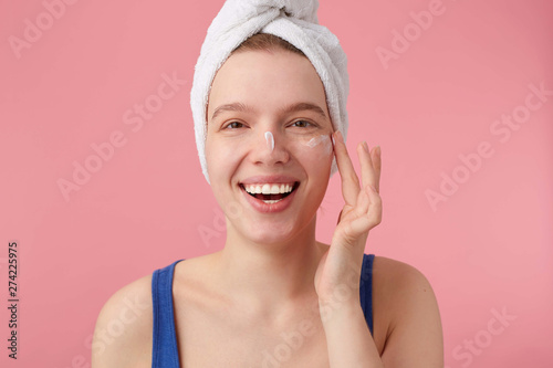 Close up of young nice smiling woman with natural beauty with a towel on her head after shower, looking at the camera over pink background and puts on face cream.