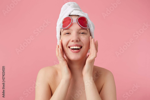 Close up of young happy woman after shower with a towel on her head, broadly smiles, touches face and smooth skin, stands over pink background.