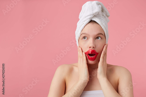 Young shocked lady after spa with a towel on her head and patch for lips, looks at the away, with wide open eyes and mouth, hears new gossip, touches cheeks with palms, stands over pink background.