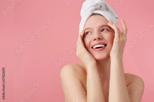 Studio photo of young happy woman after shower with a towel on her head, broadly smiles, looks away, touches face and smooth skin, stands over pink background.