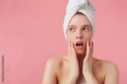 Photo of wondered young woman after shower with a towel on her head, looks away with wide open mouth and eyes, touches her cheeks, stands over pink background.