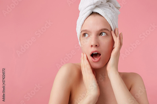 Close up of shocked young woman after shower with a towel on her head, looks away with wide open mouth and eyes, touches face, stands over pink background.