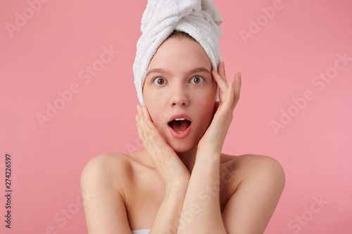 Close up of young amazed woman after shower with a towel on her head, looks at the camera with wide open mouth and eyes, touches face, stands over pink background.