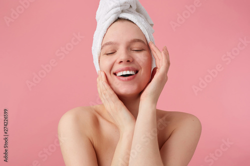 Portrait of young happy woman after shower with a towel on her head, broadly smiles with closed eyes, touches face and smooth skin, stands over pink background.