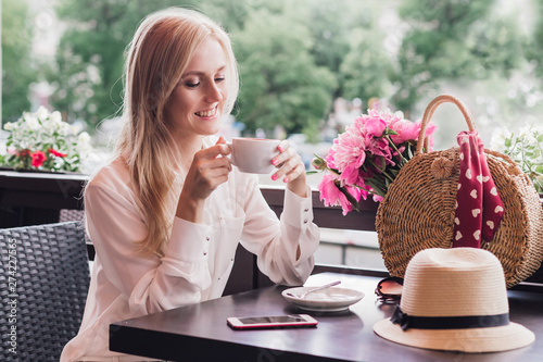 Stylish happy woman sitting in a summer cafe in her hands a cup of coffee.Wicker bag with pink flowers, a summer hat and a phone jn the table photo