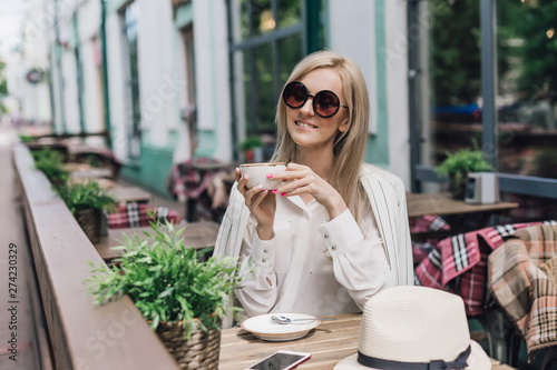 Stylish woman in sunglasses is sitting in a summer cafe in the hands of a cup of coffee
