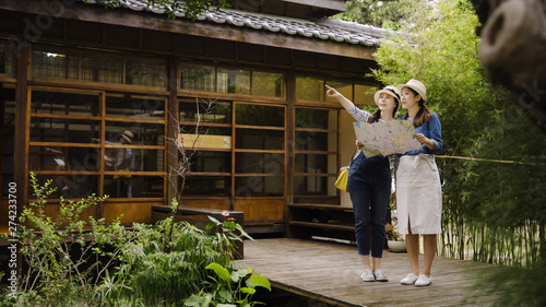 Attractive asian female traveler having conversation with best friend pointing finger discussing route. two girls searching location on paper map standing on wooden bridge out of japanese style house