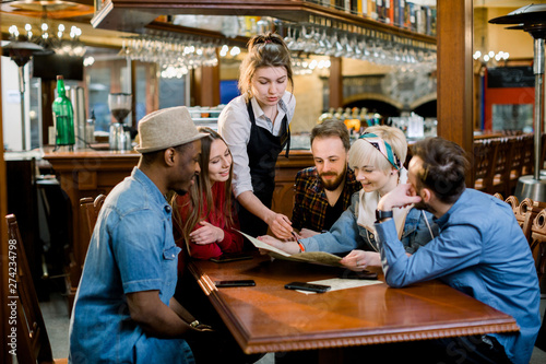 Group of diverse young friends sitting together at a table in a trendy fast food restaurant ordering food from a waitress