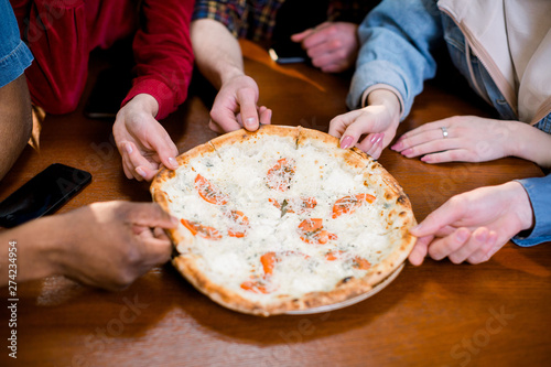Hands of diverse people taking pizza slices from wooden board dining together, multiracial friends or colleagues sharing meal having lunch in cafe restaurant