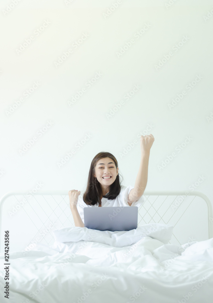 Beautiful Asian woman is smiling.Lady work with laptops on the sofa in the room in the morning.She is happy to get a new job, success, or get good news.In the white room there is a woman inside.