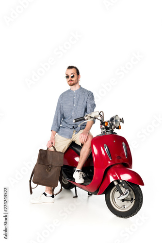full length view of handsome stylish young man sitting on red scooter and holding backpack isolated on white