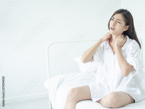 Asian women wear white pajamas in the bedroom. She has pain in the neck area.Do not focus on the main object of this image.