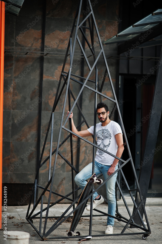 Street style arab man in eyeglasses with longboard posed inside metal pyramid construction.