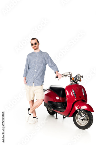 full length view of handsome young man standing near red scooter and looking at camera isolated on white