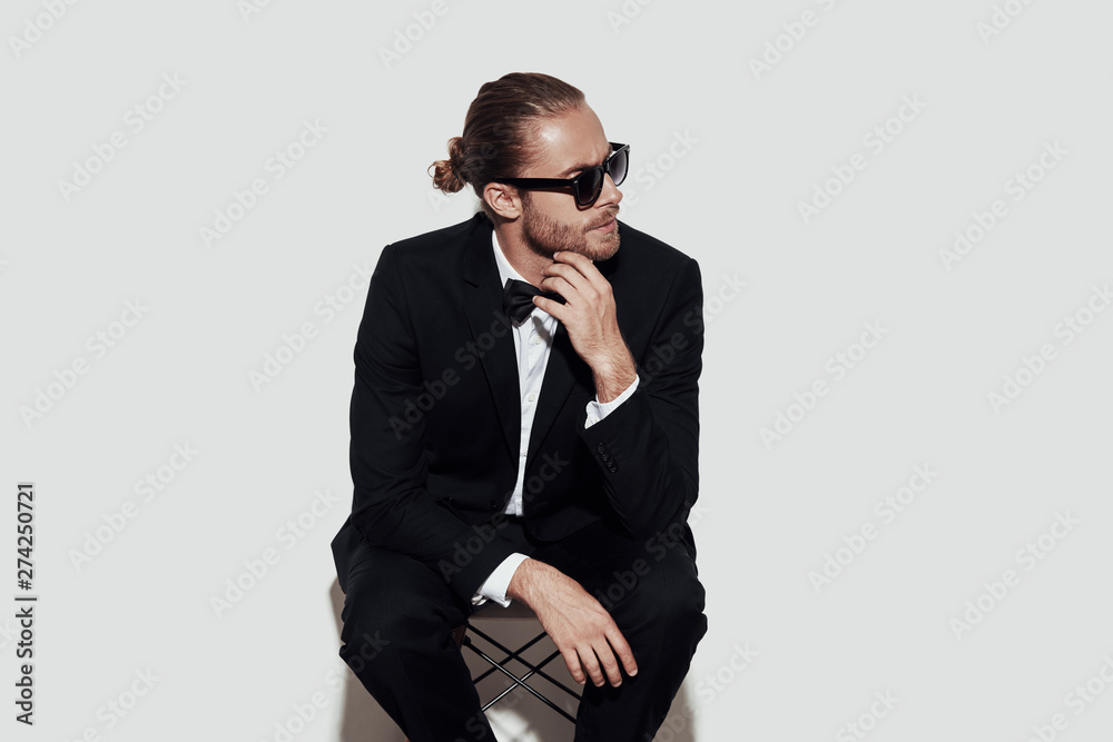 Thinking about... Charming young man in full suit keeping hand on chin and looking away while sitting against grey background Thinking about... Charming young man in full suit keeping hand on chin and looking away while sitting against grey background